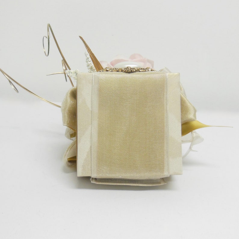 K&ouml;nnte beinhalten: Eine kleine, quadratische, beige Box mit einem zarten Blumenarrangement darauf. Die Box ist aus Stoff und hat eine Satin-Oberfl&auml;che. Die Blumen sind wei&szlig; und rosa und mit goldfarbenem Draht verziert.