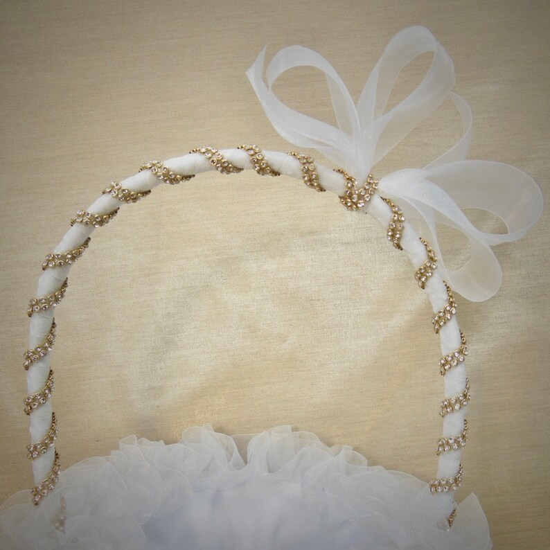 K&ouml;nnte beinhalten: Ein wei&szlig;er Blumenm&auml;dchenkorb mit einem wei&szlig;en Satinband und goldenen Strasssteinen. Der Korb ist mit einer wei&szlig;en T&uuml;llschleife verziert.