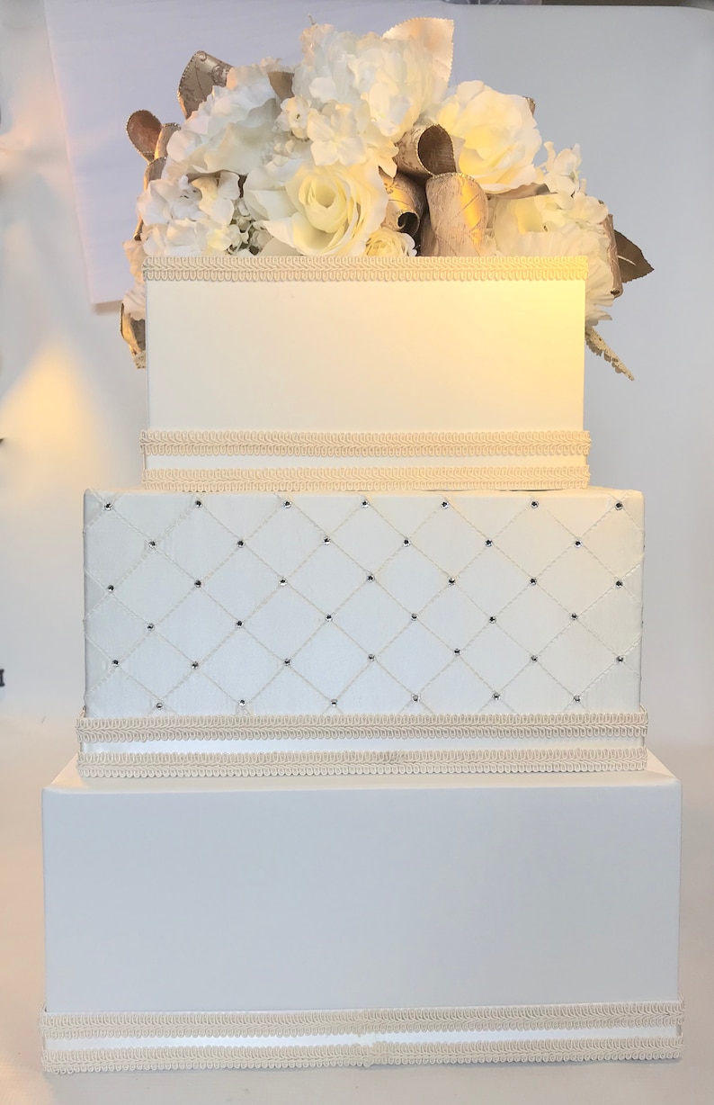 May include: A three-tiered white wedding card box with a white and gold floral arrangement on top. The middle tier has a diamond-shaped quilted pattern. Each tier has a white satin ribbon trim.