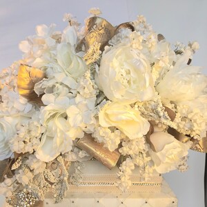May include: A white box with a white and gold floral arrangement on top. The box has a quilted pattern and a decorative trim around the top.