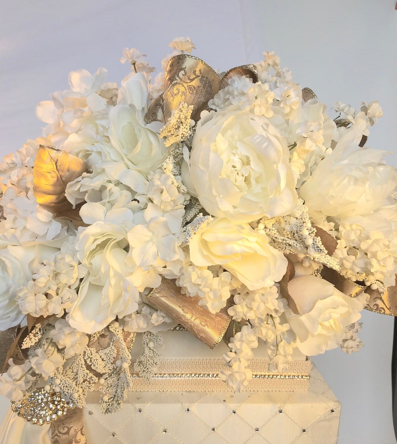 May include: A white box with a white and gold floral arrangement on top. The box has a quilted pattern and a decorative trim around the top.