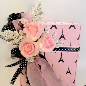K&ouml;nnte beinhalten: Eine rosa Geschenkbox mit schwarzem Eiffelturm-Muster, verziert mit rosa Rosen, wei&szlig;en Zweigen und schwarz-wei&szlig; gepunktetem Band. Eine glitzernde Brosche und ein durchsichtiges rosa Band verleihen Eleganz.