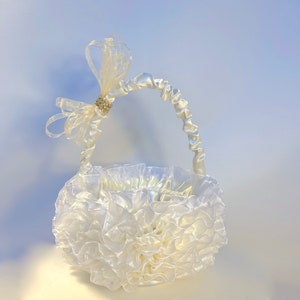 May include: A white, ruffled basket with a handle and a decorative bow. The basket is made of a soft, sheer fabric and features a sparkling rhinestone accent on the bow. The handle is also white and has a satin-like texture.