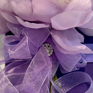 May include: A purple flower with a sparkly rhinestone embellishment is tied with a sheer purple ribbon and placed on a purple velvet box.