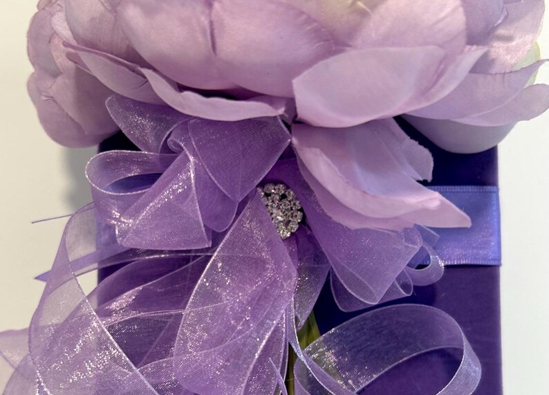May include: A purple flower with a sparkly rhinestone embellishment is tied with a sheer purple ribbon and placed on a purple velvet box.