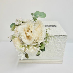 May include: White lace wedding card box with a decorative floral arrangement of white roses, baby's breath, and eucalyptus leaves. The box has a slot for cards and a rhinestone embellishment.