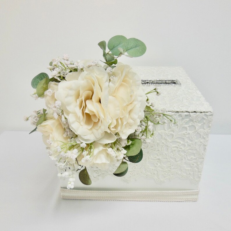 May include: White lace wedding card box with a decorative floral arrangement of white roses, baby's breath, and eucalyptus leaves. The box has a slot for cards and a rhinestone embellishment.