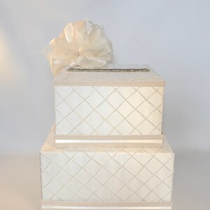 May include: A white two-tiered gift box with a diamond pattern and a white satin bow on top. The box is ideal for a wedding or other special occasion.