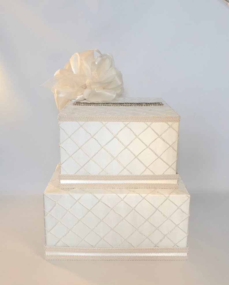 May include: A white two-tiered gift box with a diamond pattern and a white satin bow on top. The box is ideal for a wedding or other special occasion.