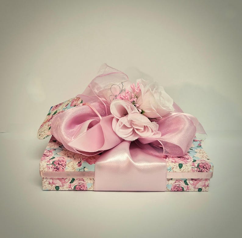 May include: A floral patterned gift box with a large pink satin ribbon and bow. The box is decorated with pink roses and light blue accents. The bow is made of sheer and satin ribbons, and topped with small pink flowers.