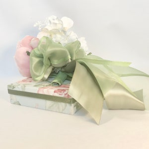 K&ouml;nnte beinhalten: Ein blasses gr&uuml;nes Satinband, das zu einer Schleife gebunden ist, um einen Strau&szlig; aus wei&szlig;en und rosa Seidenblumen. Der Strau&szlig; liegt auf einer kleinen, floral gemusterten Geschenkbox.