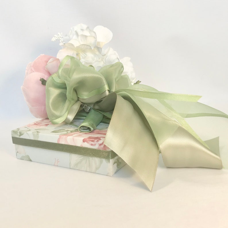 K&ouml;nnte beinhalten: Ein blasses gr&uuml;nes Satinband, das zu einer Schleife gebunden ist, um einen Strau&szlig; aus wei&szlig;en und rosa Seidenblumen. Der Strau&szlig; liegt auf einer kleinen, floral gemusterten Geschenkbox.