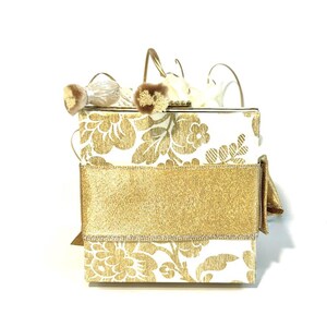 May include: A white box with a gold floral pattern and a gold ribbon. The box has a gold clasp and is decorated with white and gold feathers.
