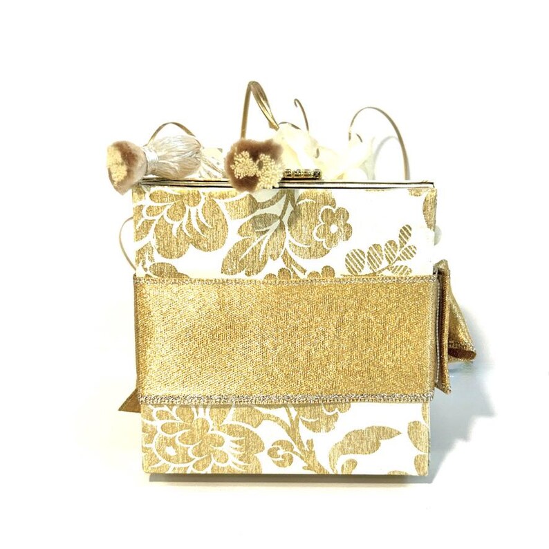 May include: A white box with a gold floral pattern and a gold ribbon. The box has a gold clasp and is decorated with white and gold feathers.