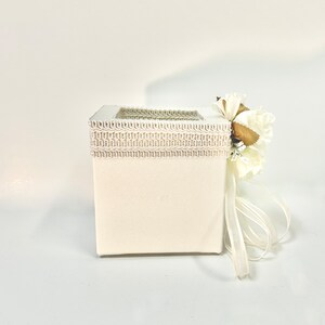 May include: A white square box with a decorative trim and a white flower arrangement on top. The box has a white ribbon tied around it.