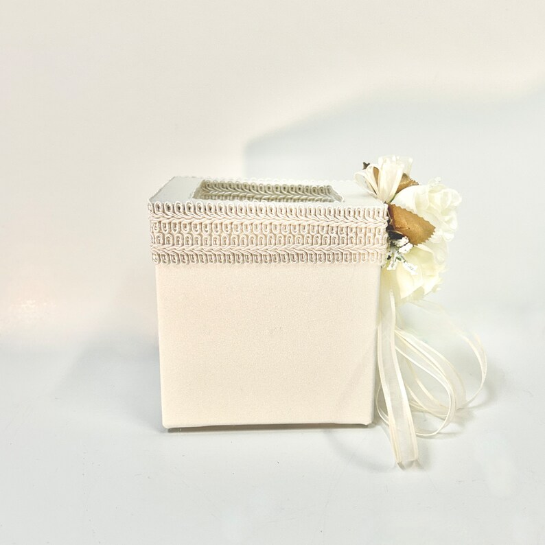 May include: A white square box with a decorative trim and a white flower arrangement on top. The box has a white ribbon tied around it.