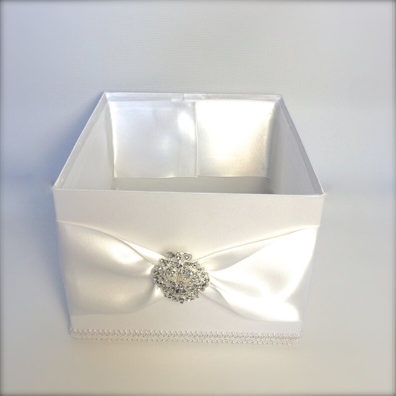 Puede incluir: Caja de regalo de sat&eacute;n blanco con un lazo de sat&eacute;n blanco y un adorno de strass. La caja tiene un borde de encaje blanco alrededor del borde inferior.