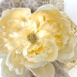 Puede incluir: Primer plano de una flor de tela de color crema con un centro de pedrer&iacute;a dorada. La flor est&aacute; sobre una cinta de estampado de leopardo beige y marr&oacute;n.