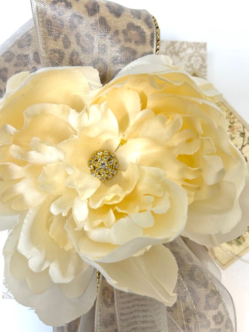 Puede incluir: Primer plano de una flor de tela de color crema con un centro de pedrer&iacute;a dorada. La flor est&aacute; sobre una cinta de estampado de leopardo beige y marr&oacute;n.