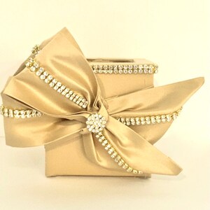 May include: A gold square box with a large satin bow and rhinestones. The bow is tied with a large rhinestone in the center.