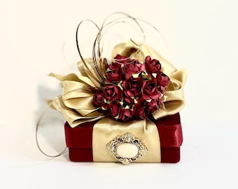 May include: A small red velvet box with a gold ribbon and a cluster of burgundy silk flowers. The box has a gold metal embellishment with a white centre.