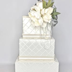 May include: A three-tiered white gift box with a diamond pattern and pearl trim. The top tier is adorned with a bouquet of white peonies and a silver ribbon.