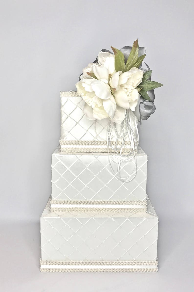 May include: A three-tiered white gift box with a diamond pattern and pearl trim. The top tier is adorned with a bouquet of white peonies and a silver ribbon.