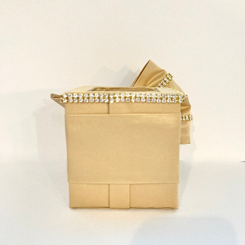 May include: A beige box with a decorative trim of rhinestones around the top edge. The box is made of fabric and has a satin finish.