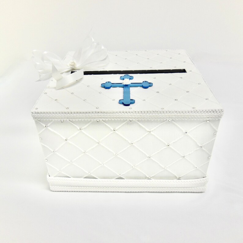 May include: White cardboard box with a blue cross on the lid. The box has a white quilted design and a white satin ribbon.