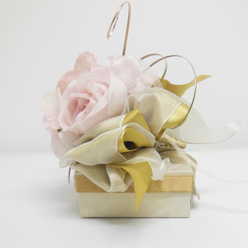 K&ouml;nnte beinhalten: Eine kleine, quadratische Geschenkbox mit einem wei&szlig;en und goldenen Design. Die Box ist mit einem Strau&szlig; rosa Rosen und einem goldenen Band verziert.