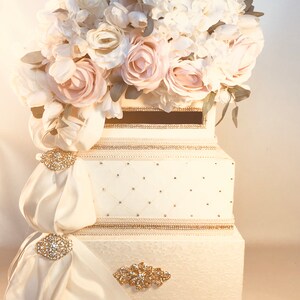 May include: An ivory-colored wedding card box with a floral arrangement of white and blush roses and hydrangeas. The box is decorated with gold trim, rhinestones, and satin drapes with jeweled brooches.