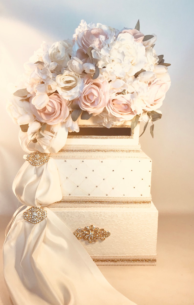 May include: An ivory-colored wedding card box with a floral arrangement of white and blush roses and hydrangeas. The box is decorated with gold trim, rhinestones, and satin drapes with jeweled brooches.