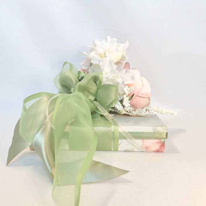 K&ouml;nnte beinhalten: Eine gr&uuml;ne Geschenkbox mit einem gr&uuml;nen Satinband und einem Strau&szlig; wei&szlig;er und rosa Blumen darauf.