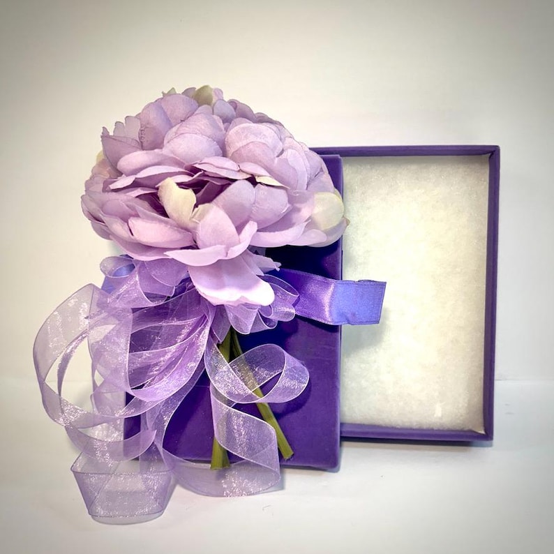 May include: A purple gift box with a lavender silk flower arrangement tied with a sheer purple ribbon. The box is open and the inside is lined with white.