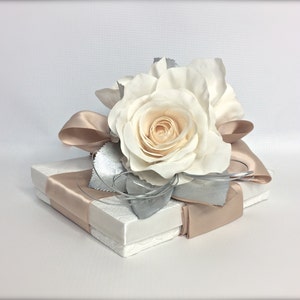May include: A white gift box with a satin ribbon and a single white rose on top. The rose is accented with silver leaves.