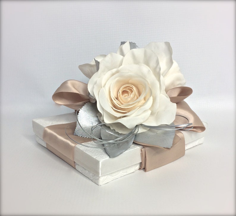 May include: A white gift box with a satin ribbon and a single white rose on top. The rose is accented with silver leaves.