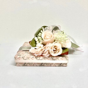 May include: A small, rectangular box wrapped in a floral patterned paper with a light green ribbon and a bouquet of white roses and greenery on top.