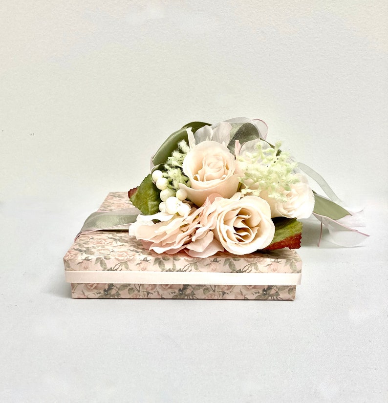 May include: A small, rectangular box wrapped in a floral patterned paper with a light green ribbon and a bouquet of white roses and greenery on top.