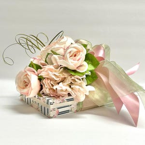 K&ouml;nnte beinhalten: Eine dekorative Geschenkbox mit einem Bouquet aus hellrosa Rosen und Hortensien. Die Box ist mit einem durchsichtigen gr&uuml;nen und rosa Band umwickelt, und die Box selbst ist mit einem Blumenmuster verziert. Gr&uuml;ne Bl&auml;tter und lockige gr&uuml;ne Stiele erg&auml;nzen das Blumenarrangement.