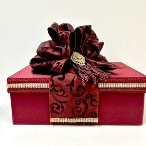 May include: A burgundy gift box with a large, ornate bow. The bow is embellished with a sparkling brooch and tassel. The box is trimmed with a rhinestone border and a patterned burgundy fabric.
