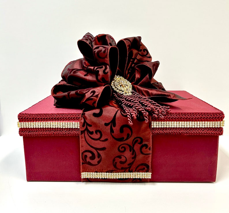 May include: A burgundy gift box with a large, ornate bow. The bow is embellished with a sparkling brooch and tassel. The box is trimmed with a rhinestone border and a patterned burgundy fabric.