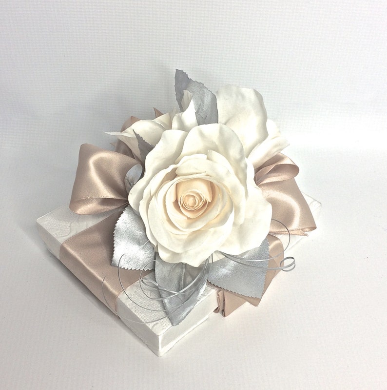 May include: A white gift box with a beige satin ribbon and a white rose with silver leaves.