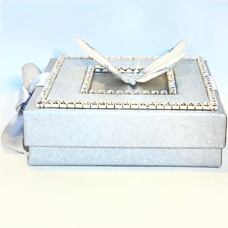 May include: A silver gift box with a white butterfly on top. The box is decorated with a border of rhinestones.