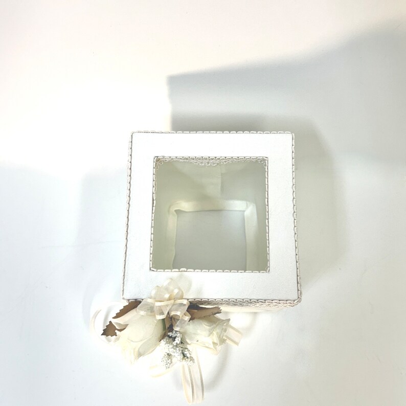 May include: White square tissue box with a lace design and a clear window. The box is decorated with a white flower and ribbon.