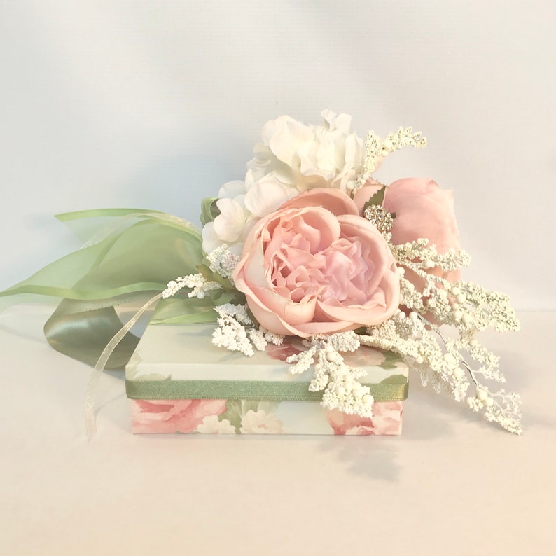 K&ouml;nnte beinhalten: Eine kleine, florale Geschenkbox mit einem gr&uuml;nen Band und einem Strau&szlig; aus rosa und wei&szlig;en Blumen. Die Blumen sind in einer Traube angeordnet und beinhalten eine gro&szlig;e rosa Rose, wei&szlig;e Hortensien und wei&szlig;e Zweige.
