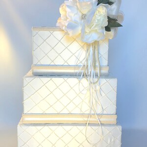 May include: Three white boxes with a diamond pattern and a white flower arrangement on top. The boxes are stacked on top of each other and have a cream colored trim.