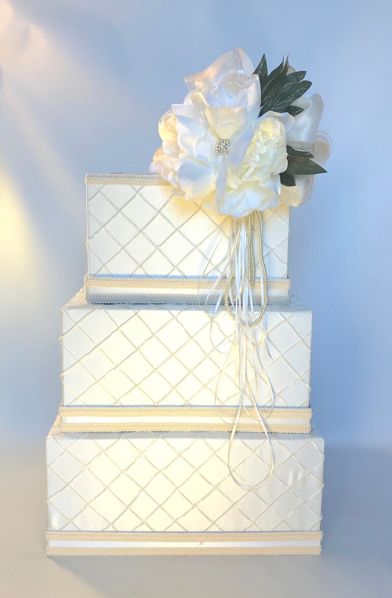 May include: Three white boxes with a diamond pattern and a white flower arrangement on top. The boxes are stacked on top of each other and have a cream colored trim.
