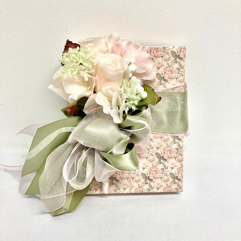 May include: A small, square box wrapped in a floral patterned paper with a green ribbon and a bouquet of white and pink flowers.