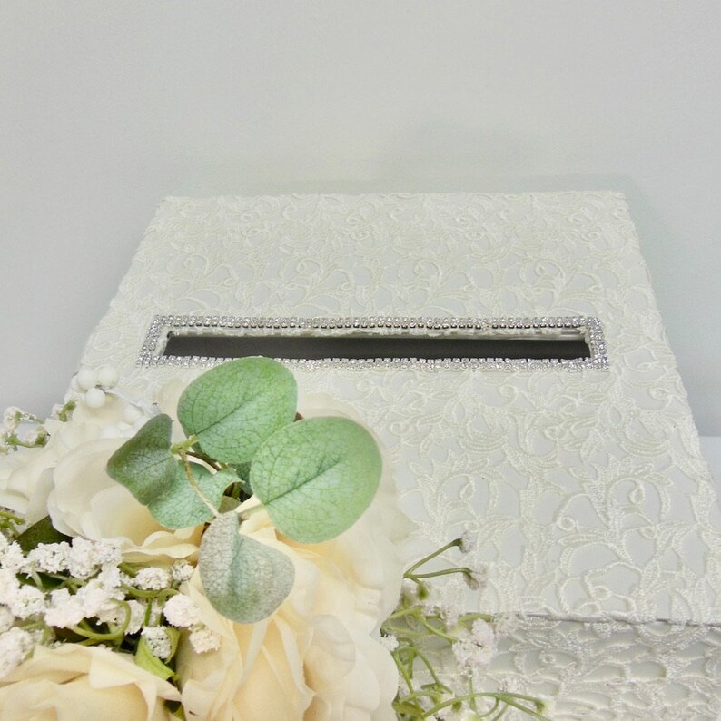 May include: White lace wedding card box with a silver rhinestone trim around the opening. The box is decorated with a white floral bouquet.