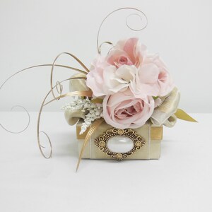K&ouml;nnte beinhalten: Eine kleine, quadratische, beige Box mit goldener Verzierung und einer dekorativen goldenen Brosche mit einer Perle in der Mitte. Die Box ist mit einem Strau&szlig; rosa Rosen und wei&szlig;en Blumen verziert.